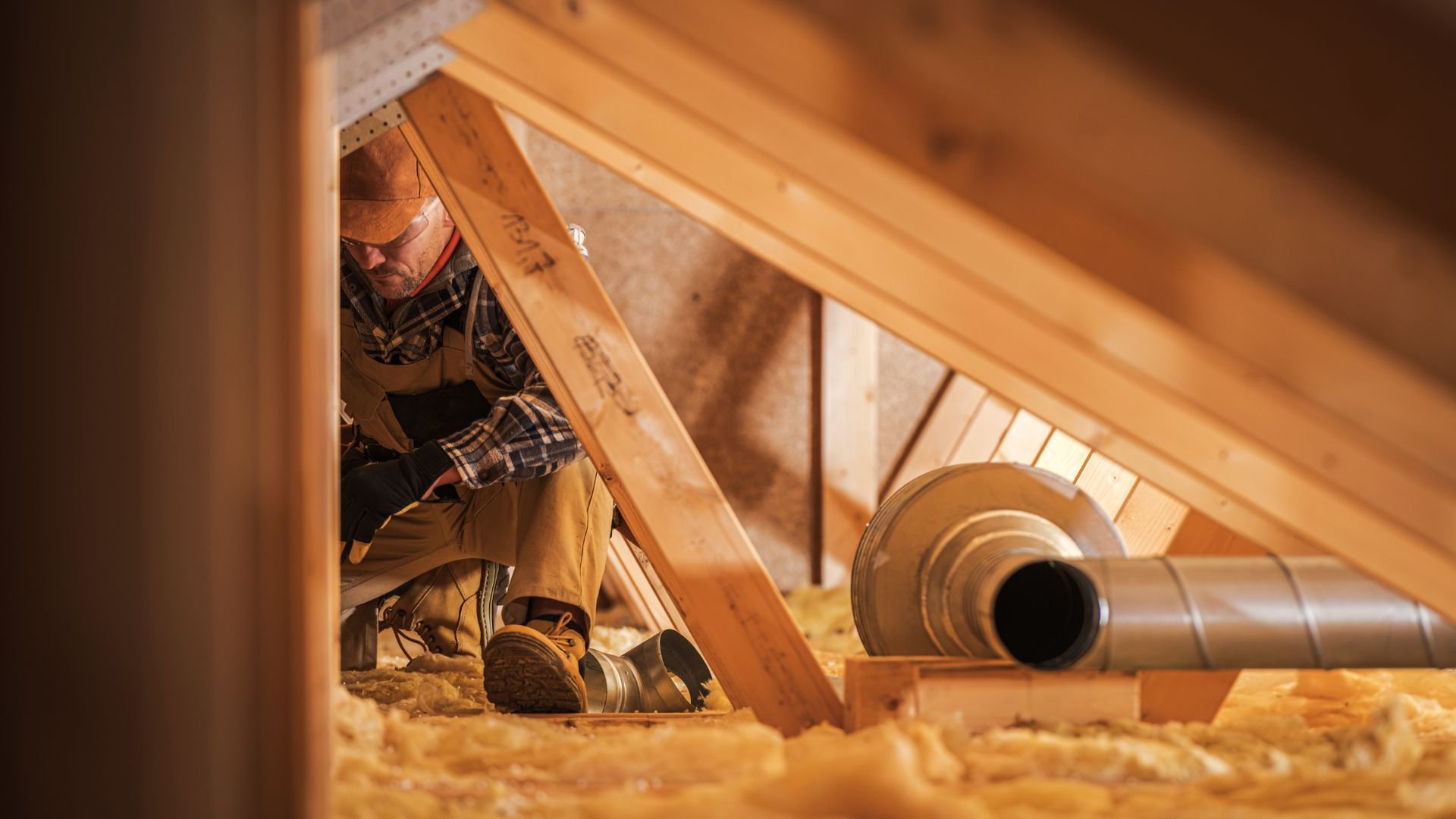 Worker installing ductwork in wooden attic with insulation and metal pipes